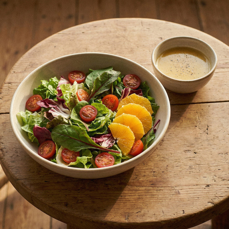 Salad with greens, tomatoes, and oranges in a bowl on a wooden table with a side of dressing.