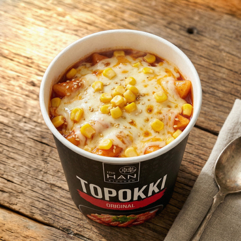 Topokki in a cup with cheese and corn on a wooden table, branded as 'The Han Kitchen'.